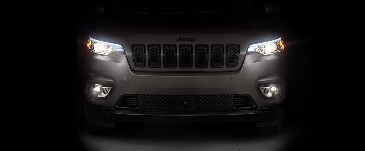 The front end of a gray 2023 Jeep Cherokee Altitude Lux with its headlamps and fog lamps lit.