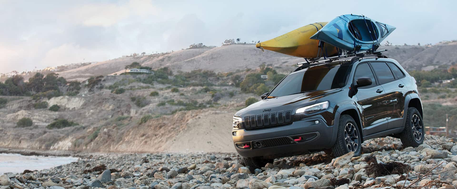 A 2023 Jeep Cherokee Trailhawk with two kayaks strapped to its roof rack, crawling over stones on a rocky beach.