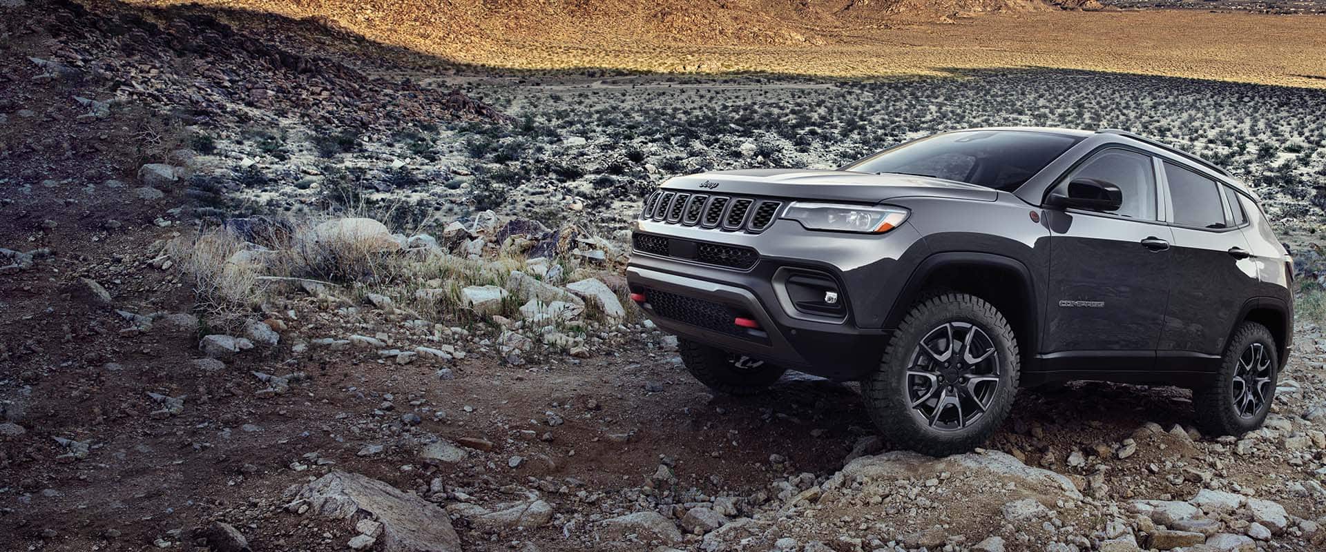 A gray 2024 Jeep Compass Trailhawk crawling over large and small rocks and stones off-road.